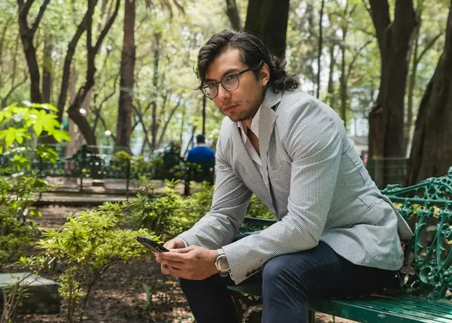 A man in a blazer on a park bench, checking his phone.